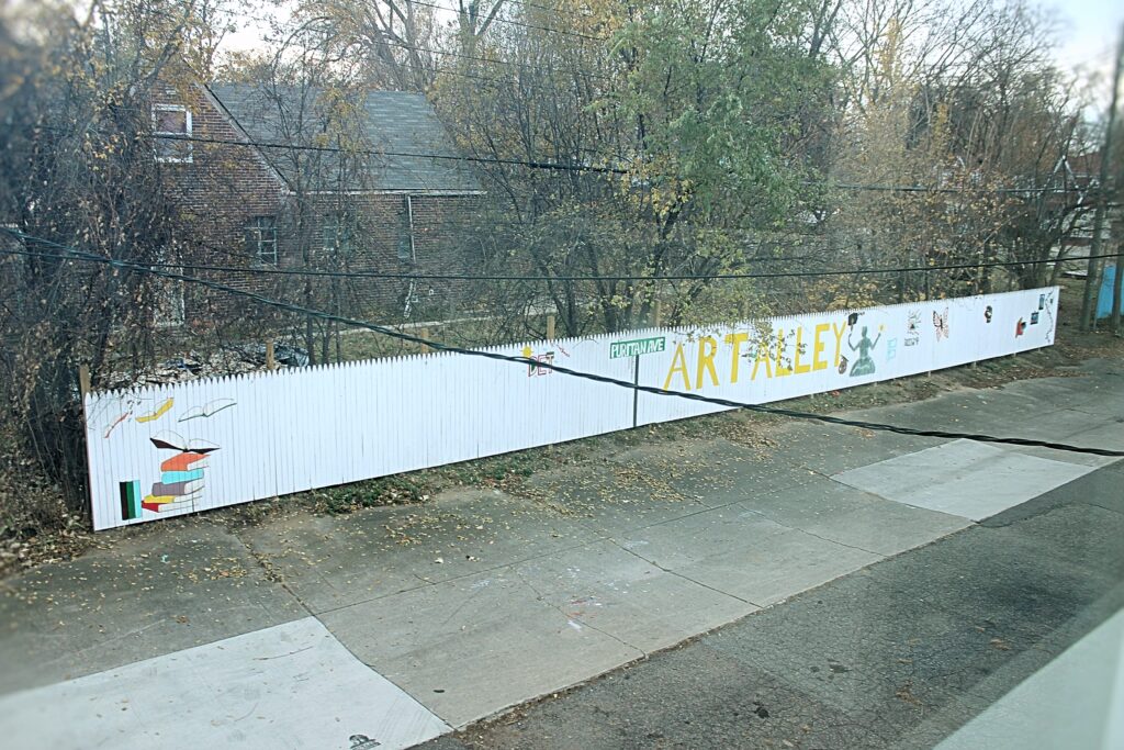 Looking down across the street from a white-painted picket fence reading "Puritan Ave ART ALLEY".