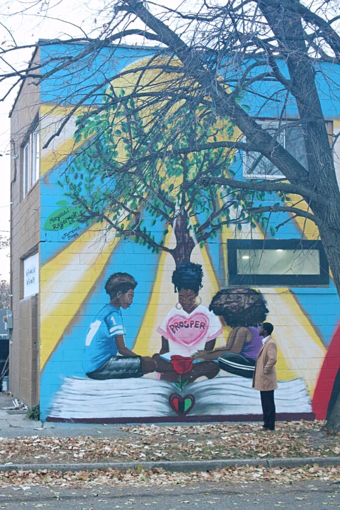 Mural of 3 black children sitting on an opened book under a tree, with the word "Prosper" inside a pink heart floating in front of them.