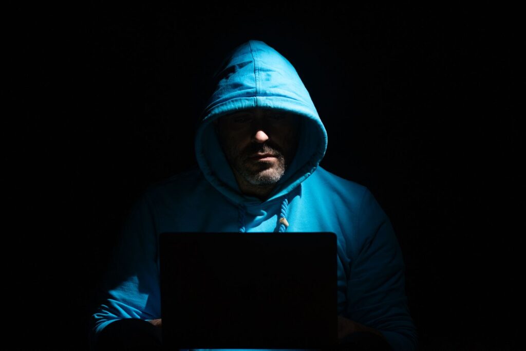 SCAM ALERT - Gmail Addresses Impersonating DBF Team Image 1 A man in a blue hoodie focused on a laptop in a dark room, conveying secrecy and intrigue.